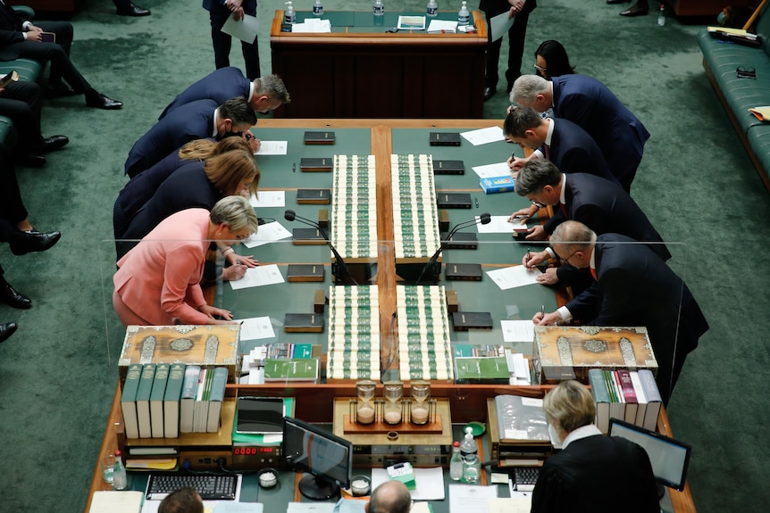 ten cabinet ministers stand around the dispatch box, bent over signing documents.