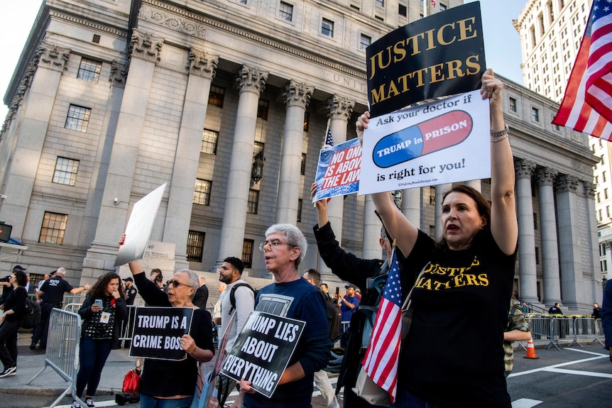 A group of protesters holding posters criticising Donald Trump, on a street outside a large government building near metal fence