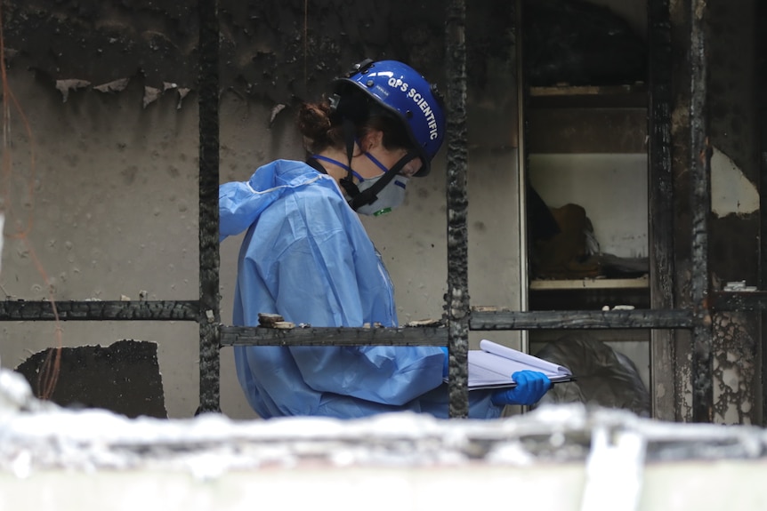 A forensic officer in a mask, helmet and PPE looks down at a clip board in the middle of a burnt out house.