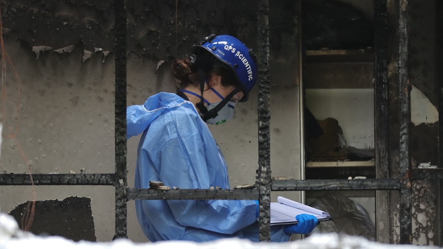 A forensic officer in a mask, helmet and PPE looks down at a clip board in the middle of a burnt out house.