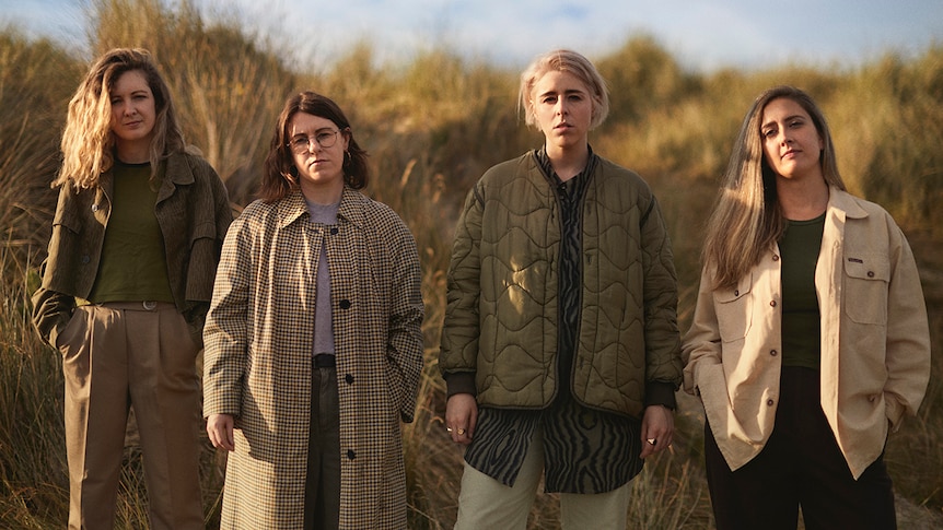 Four women stand side by side in a field wearing coats, looking at the camera