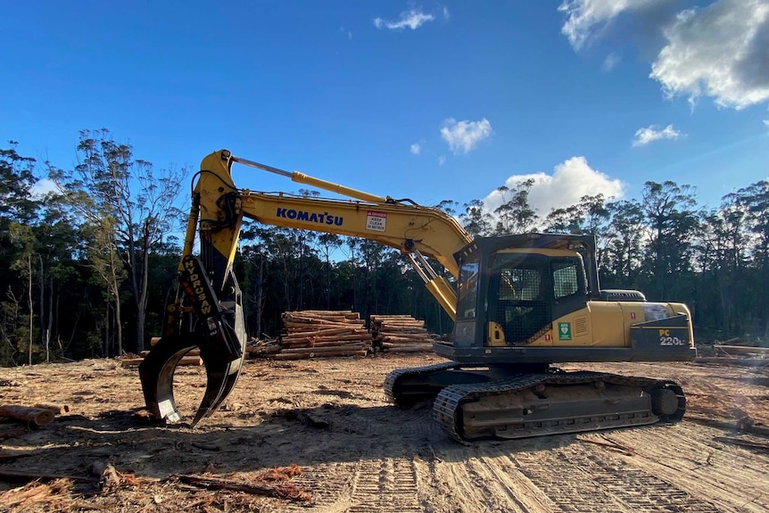 A yellow digger with timber logs in the background.