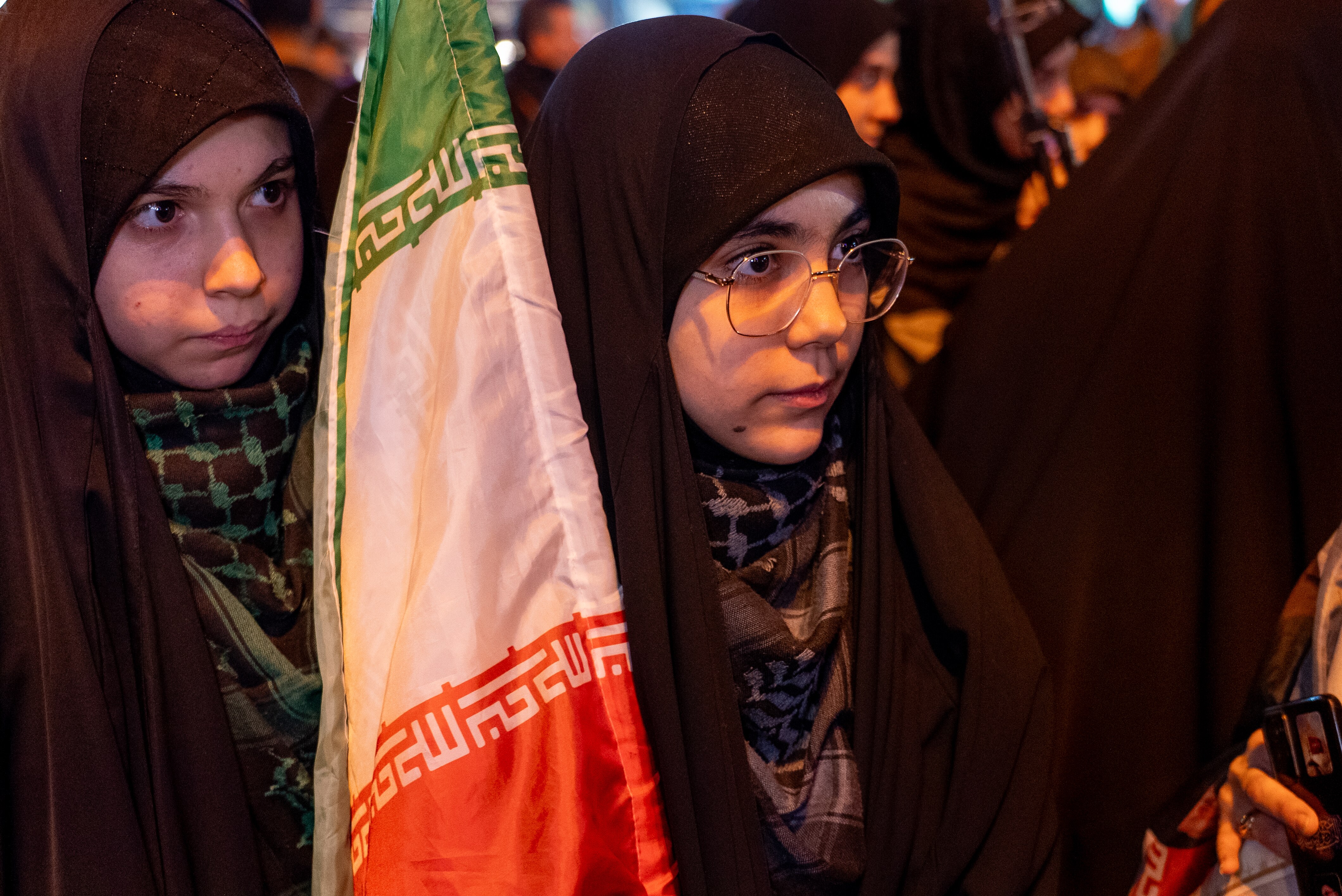 A teenage girl wearing traditional Islamic attire holding the Iranian flag beside her.