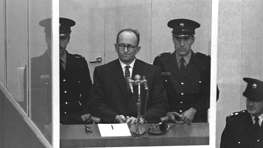 Black and white photograph of Adolf Eichmann standing flanked by two guards in a protective glass booth