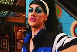 View through a car window as dark-haired drag queen leans on the door giving a dirty look and waitress smiles in background.