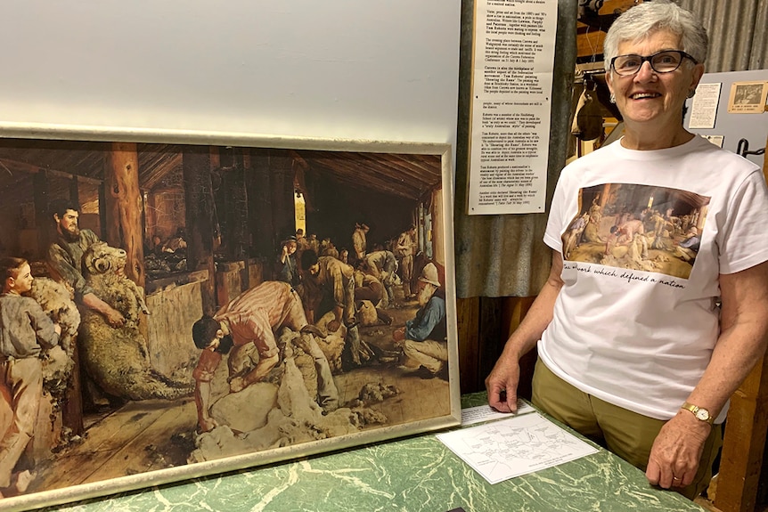 An elderly woman with grey hair and glasses smiles next to a painting depicting shearers in a shed with sheep.