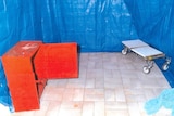 A photo of a room with the walls covered in blue tarpaulin, with a wood chest on the left and trolley on the right.