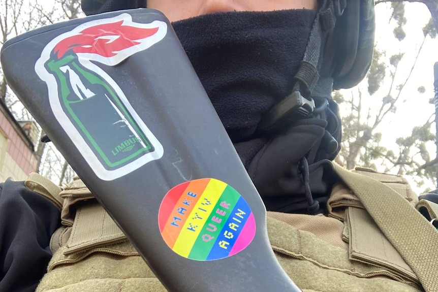 A man in military gear holding a rifle with a molotov cocktail sticker and a rainbow sticker reading "Make Kyiv Queer Again". 