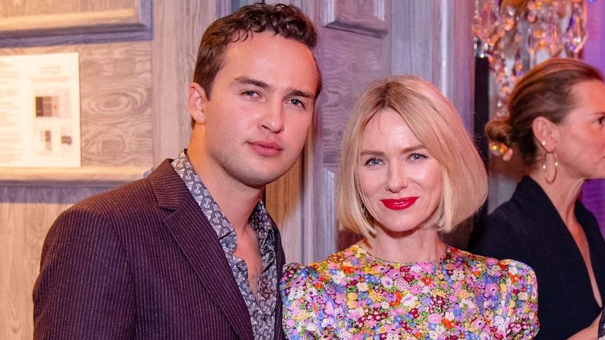 Mojean Aria, wearing a maroon striped jacket and floral shirt, stands with Naomi Watts in a floral dress.