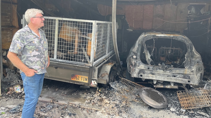 A man standing in a garage with a burnt-out trailer and car