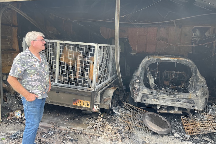 A man standing in a garage with a burnt-out trailer and car
