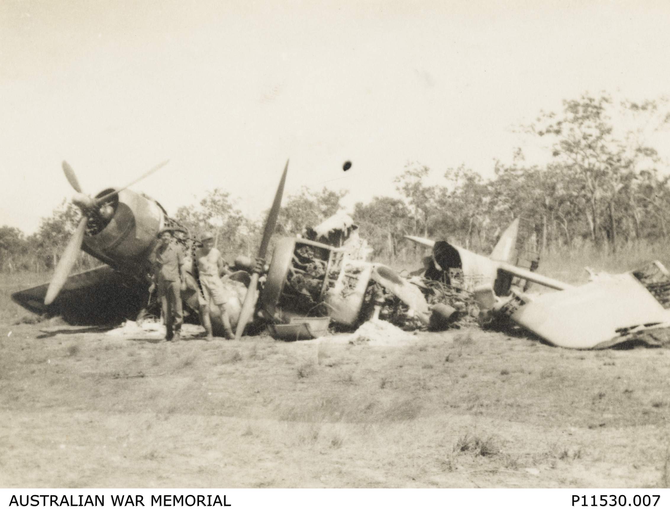 A historical, black and white photo showing two men in a field in front of a destroyedWWII-era plane.