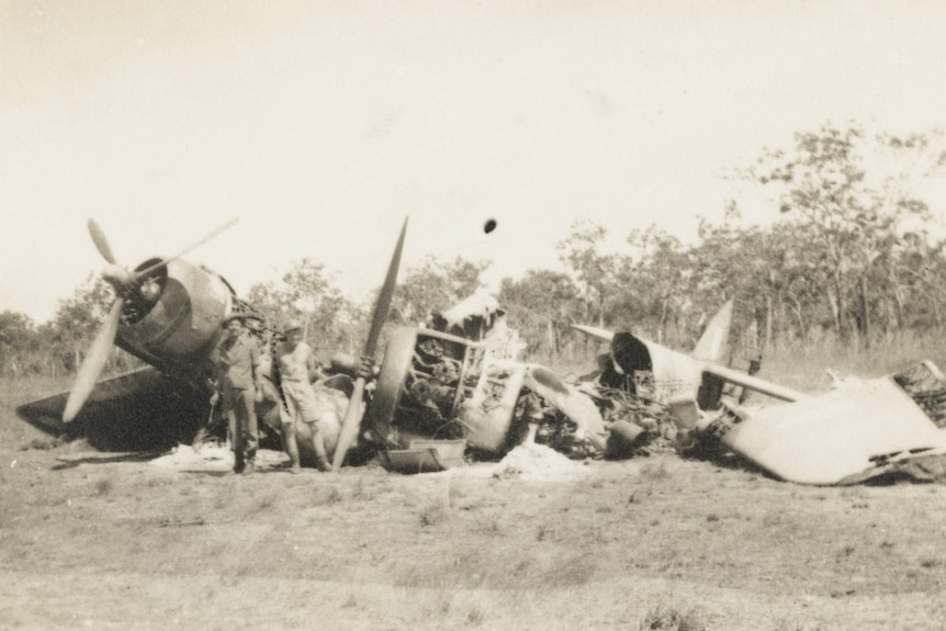 A historical, black and white photo showing two men in a field in front of a destroyedWWII-era plane.