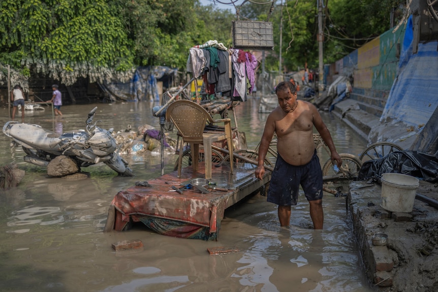 A man without a shirt stands in muddy floodwater next to furniture covered in silt.