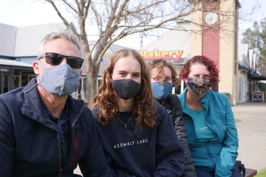 A picture of a family wearing masks sitting on a bench with a tree in the background.