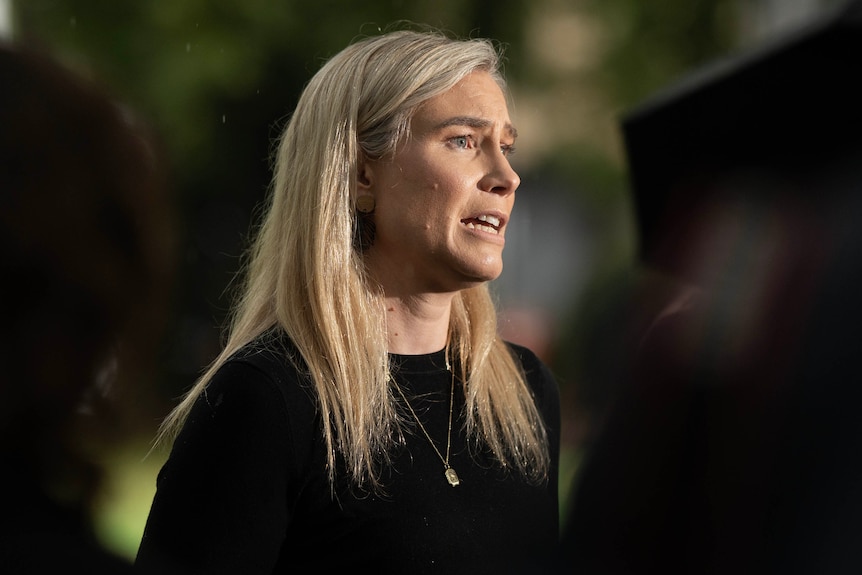 A woman speaks in front of media underneath trees.