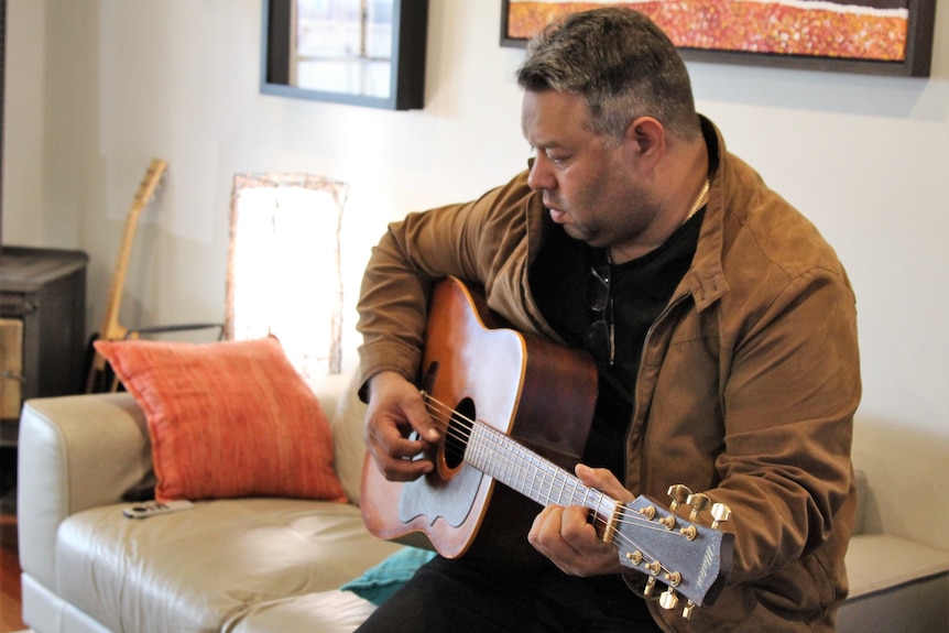 An Indigenous man sits on a cream couch and plays guitar 
