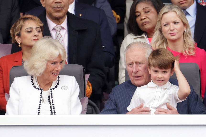 Prince Louis sits atop Charles's knee, scratching his head while the monach holds him. Camilla, seated next to them smiles