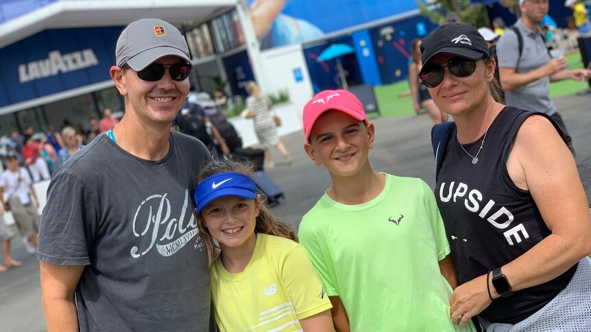 A family of four stand outside Rod Laver Arena all wearing caps on a sunny day.