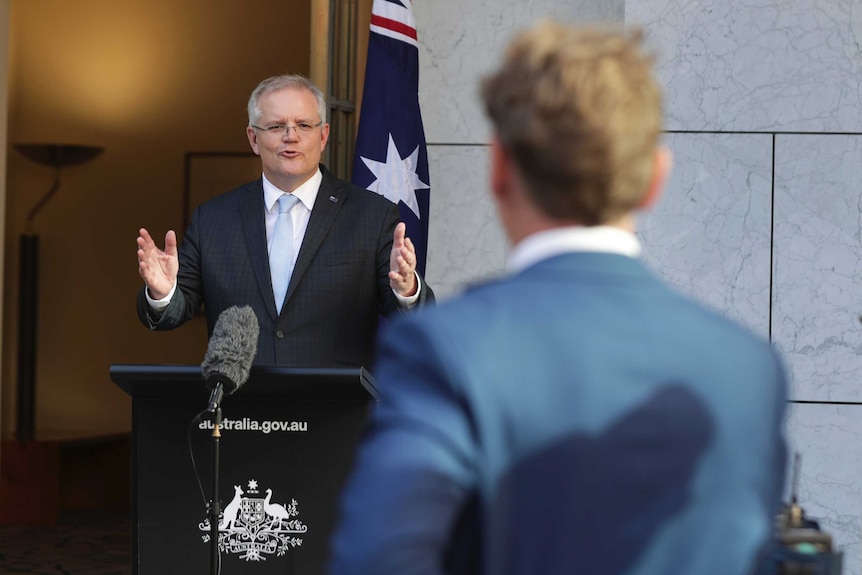 Back of Probyn's head looking towards Scott Morrison standing behind podium in front of microphone.