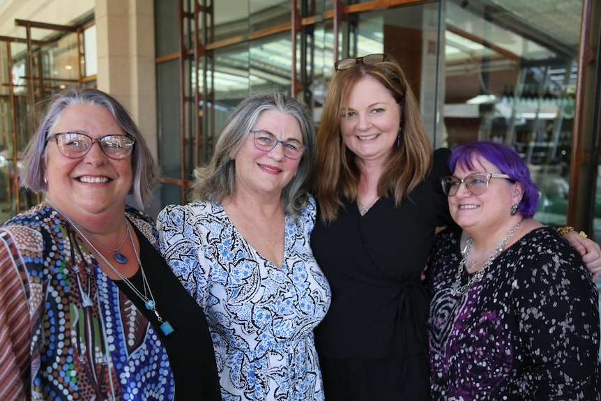 Four women standing in front of large glass doors with arms around each other, each smiling widely.