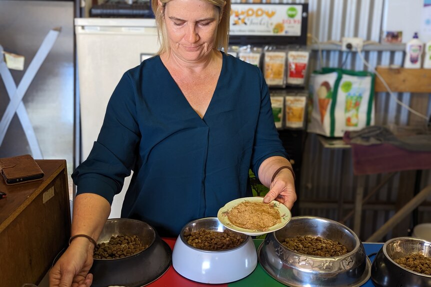 A white woman, Melissa, with blonde hair sprinkles dehydrated meat flakes on dog kibble in metal bowls in her shed.