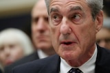 Robert Mueller looks to the left with his mouth open and his eyes wide. He wears a suit and people are seen behind him.