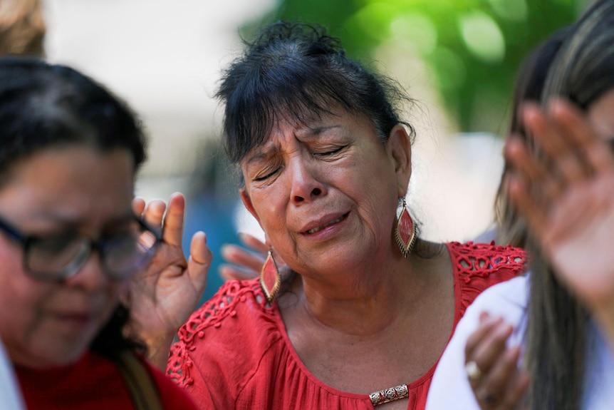 A middle-aged woman holds one hand up in prayer as she sobs in a crowd of people