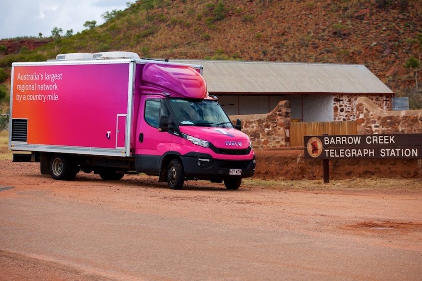 A bright pink Tesltra van dirving past a sign that says "Barrow Creek Telegraph Station".