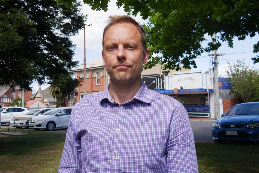 A man in a checked shirt stands in front of a cars parked along a street. 