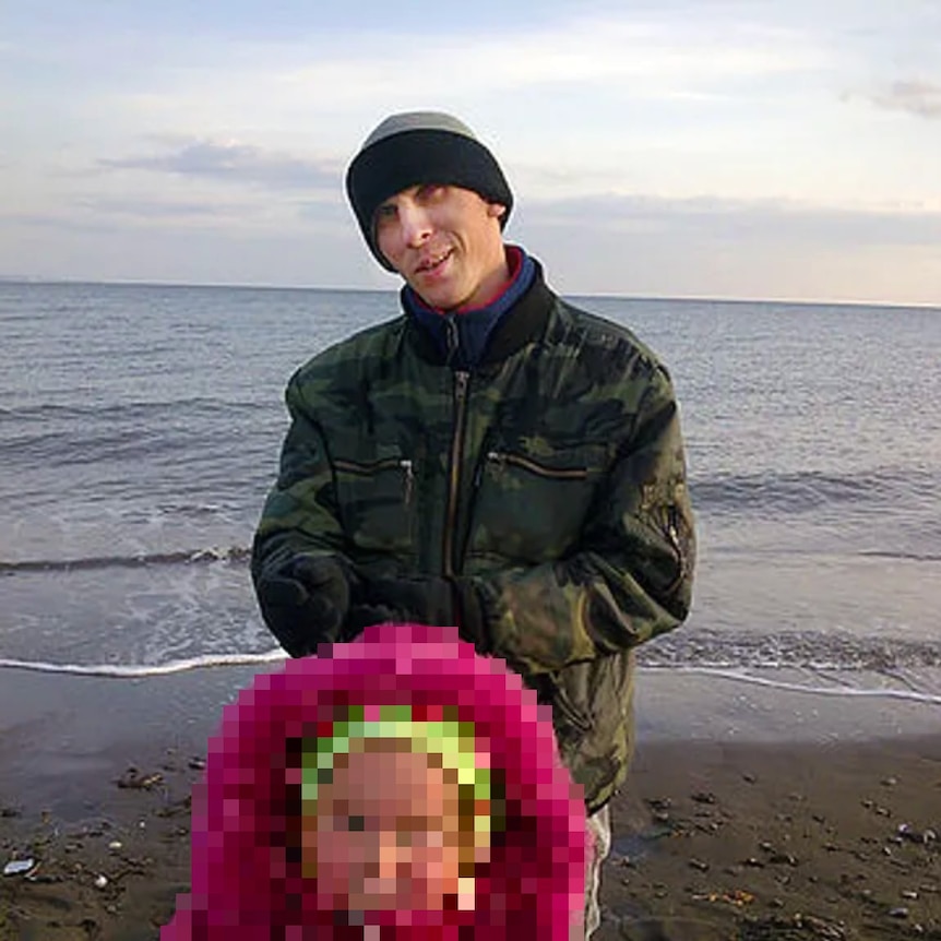 A man wearing a beanie and standing on a beach smiles at the camera. A small child with its face blurred stands in front of him