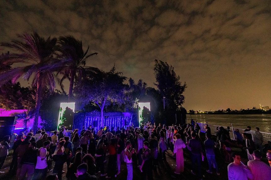 people dance outside in front of a stage near a river and large trees at night