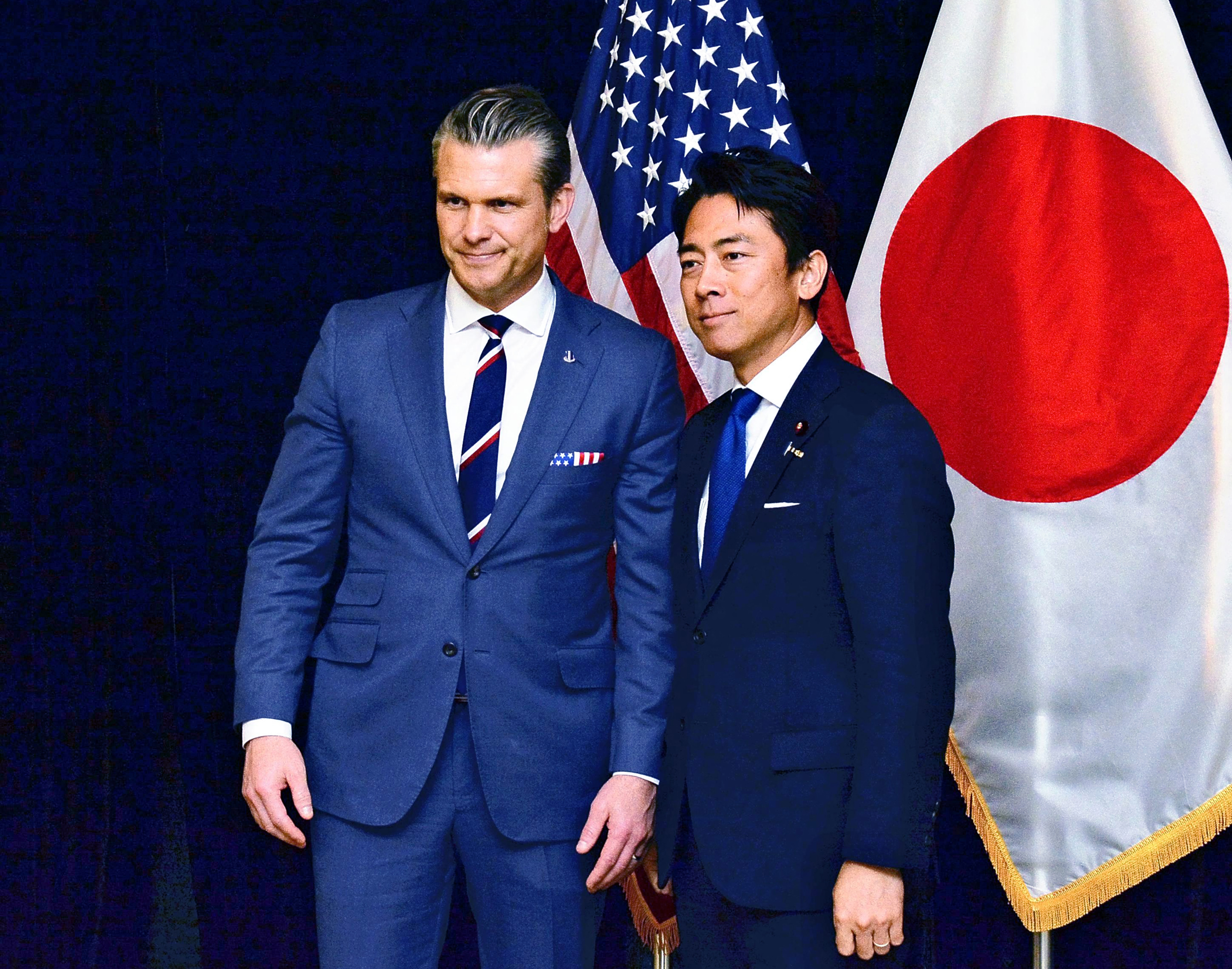 Dos hombres trajeados posan juntos frente a una bandera estadounidense y otra japonesa.