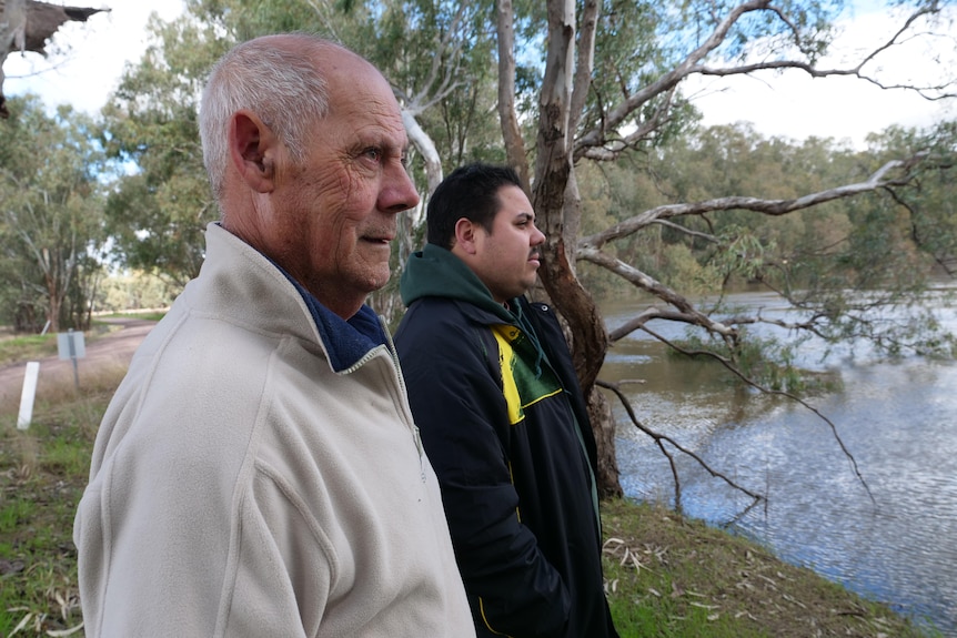 A serious older balding man with grey hair and a young man stand side by side looking out towards a river.