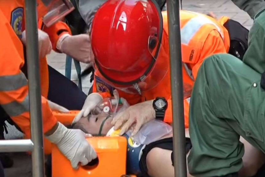 A group of people in bright orange protective gear gather around a man laying on the pavement with an oxygen mask on his face.