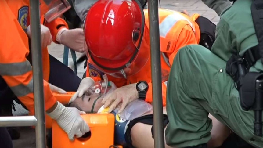 A group of people in bright orange protective gear gather around a man laying on the pavement with an oxygen mask on his face.