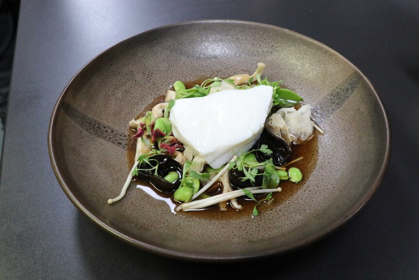 A brown plate is photographed with a dish of toothfish, mushrooms, beans and ginger on a dark table.