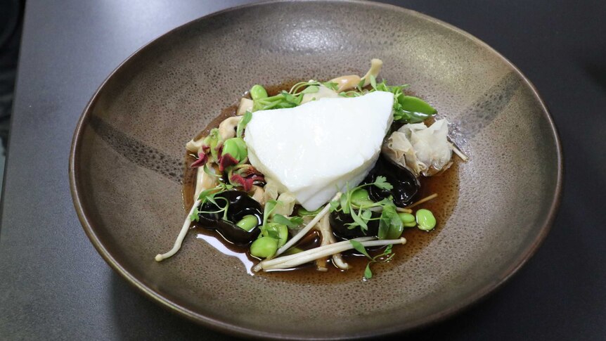 A brown plate is photographed with a dish of toothfish, mushrooms, beans and ginger on a dark table.