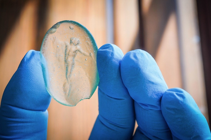 A hand wearing blue gloves holds a small glass disc with an image of a body inside it.