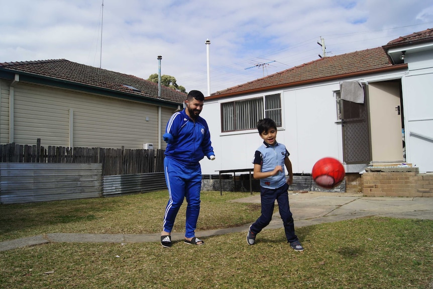 A man who has both arms missing plays soccer in a backyard with his son in a suburban yard