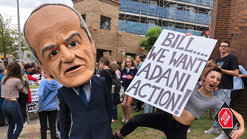 A man with a giant Bill Shorten costume head is kicked by a protester holding a sign that reads 'Bill...we want Adani action'