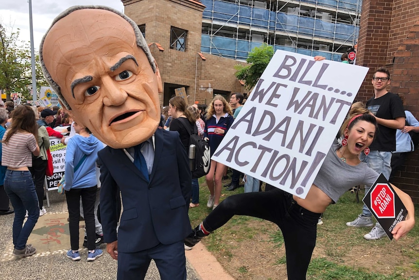 A man with a giant Bill Shorten costume head is kicked by a protester holding a sign that reads 'Bill...we want Adani action'