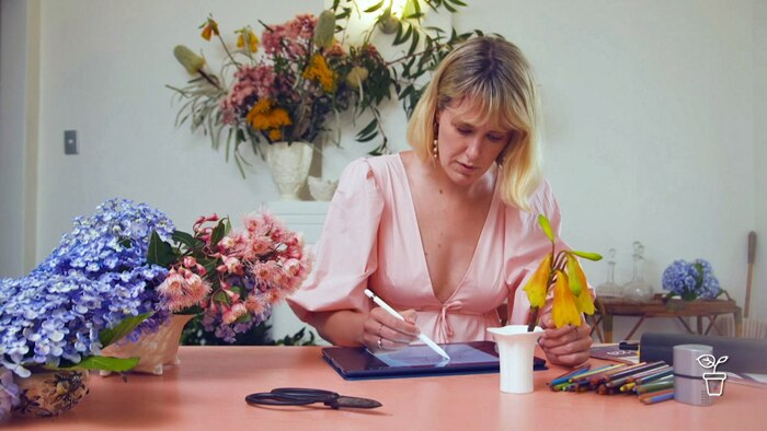 Woman sitting at a desk with vases of flowers drawing on an electronic tablet