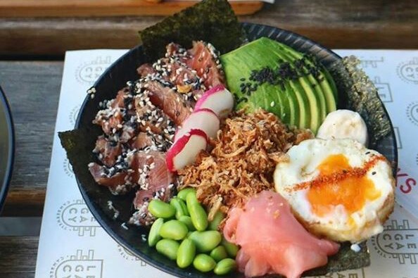 A poke bowl of avocado, egg, seared beef, radish and pickled ginger is artistically laid out on a wooden table.