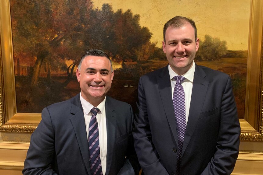 Two men smile in front of a painting.