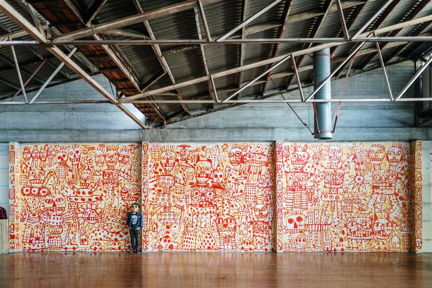 A wide shot of a man standing in front of a big colourful mural in a warehouse