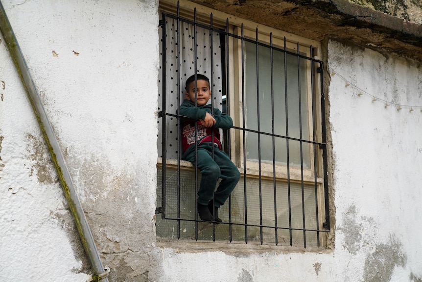 A young boy sitting in a window, behind protective bars.