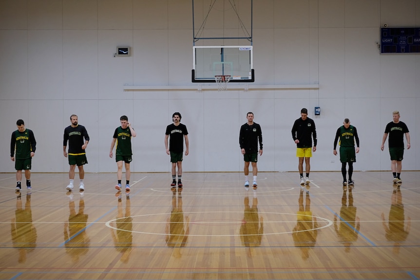 Four men walk in a line under a basketball ring on an indoor court. They wear Australian training uniforms.