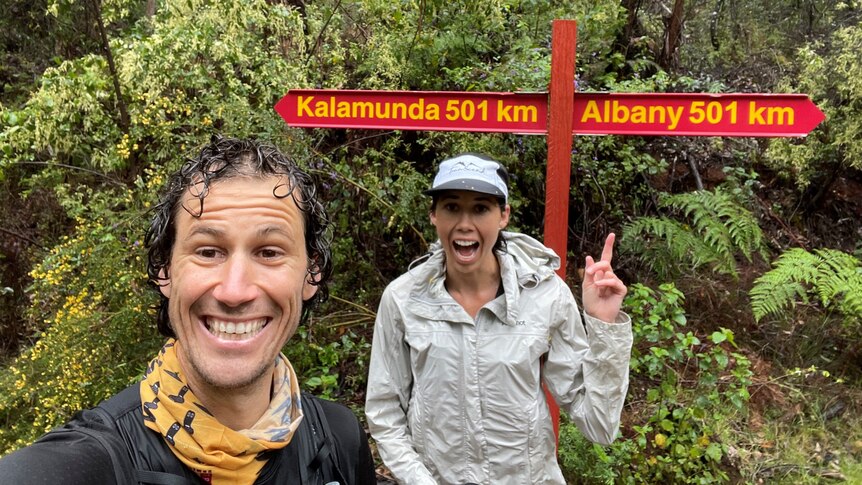 two people with trees behind them, smile and point to a sign post that says Kalamunda in one direction and Albany in the other 