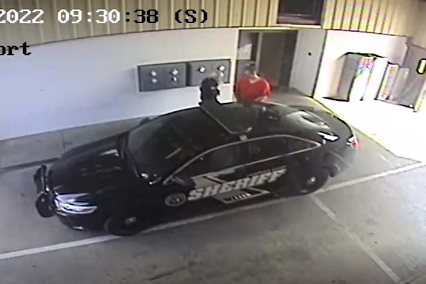 A blonde woman in uniform opens the door of a black car bearing large sheriff insignia on the side, for a man in orange